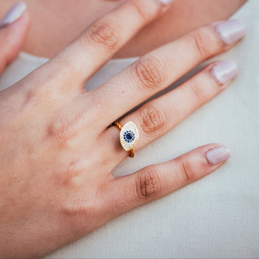 Close-up of a hand wearing an eye-shaped ring with a blue gemstone, with a necklace and earrings in the background.