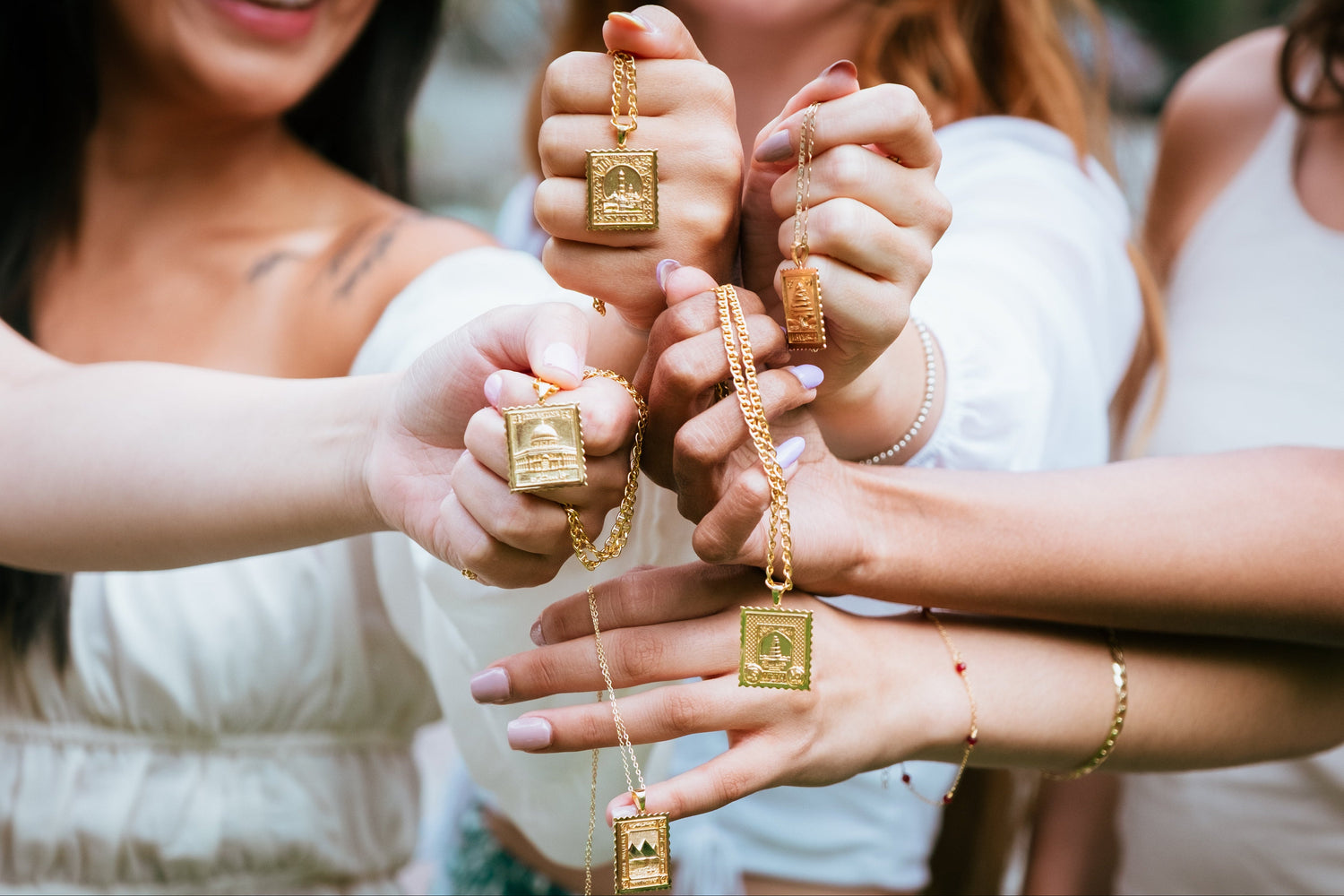 Three women holding gold necklaces with square pendants in a close-up shot.