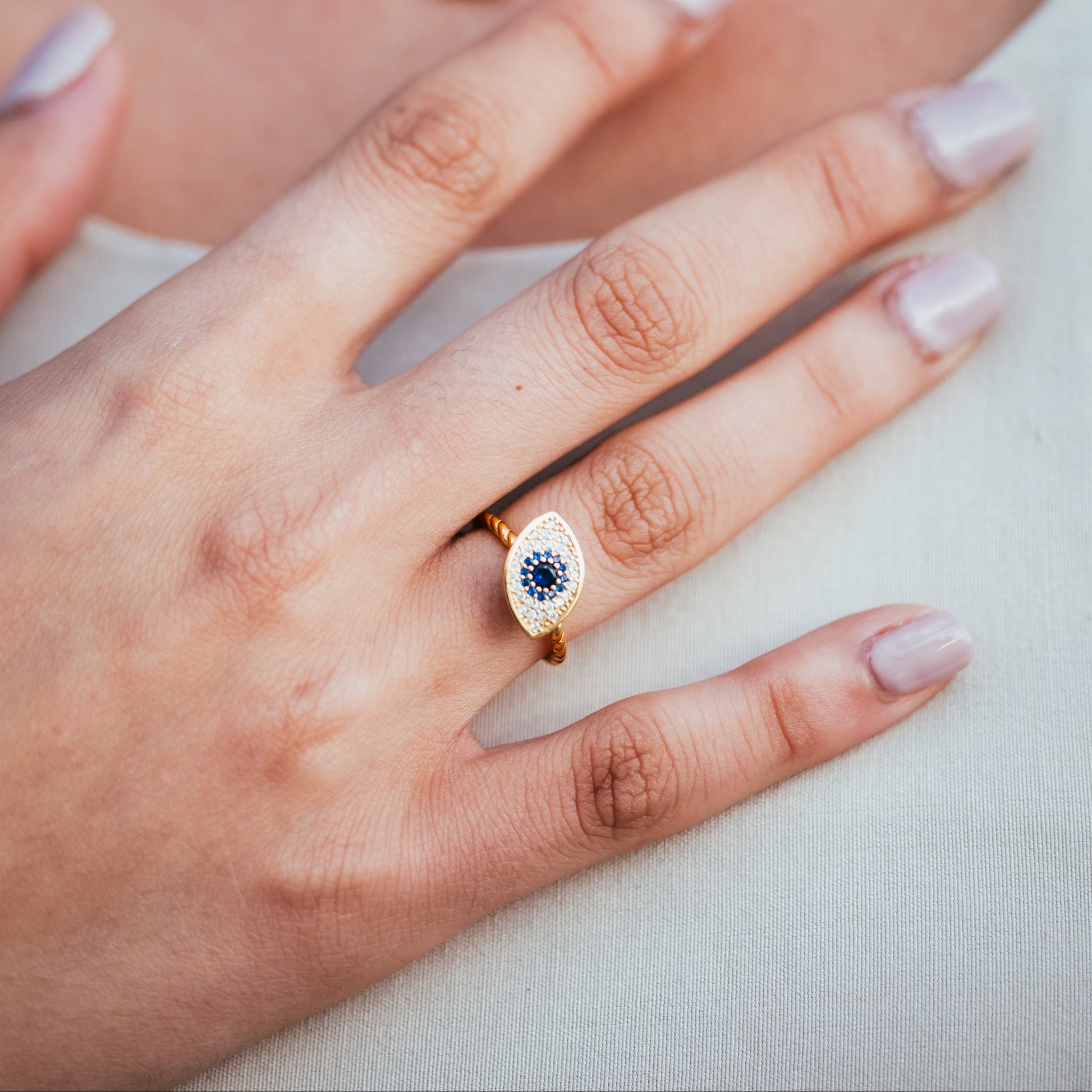 Close-up of a hand wearing an eye-shaped ring with a blue gemstone, with a necklace and earrings in the background.