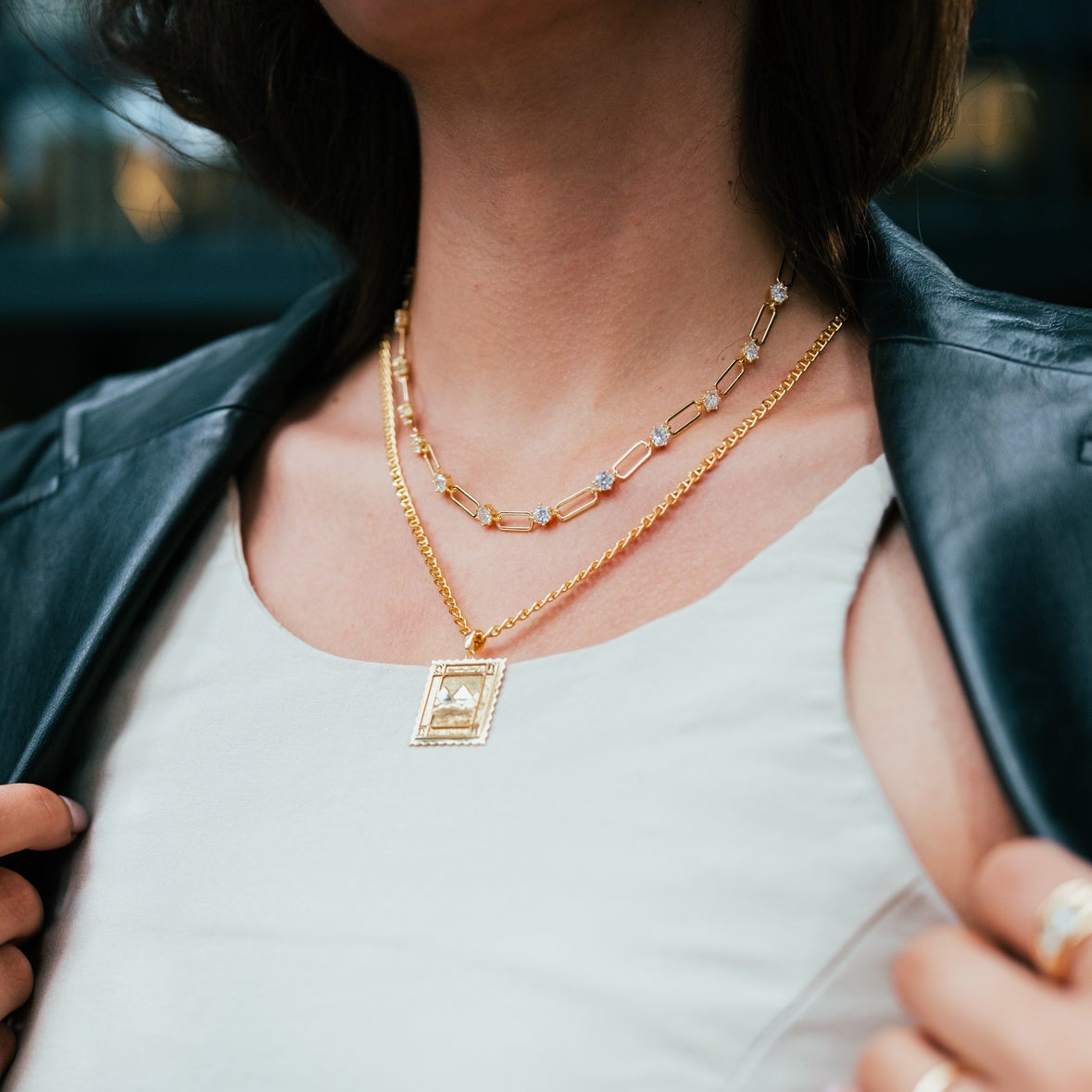 Person wearing a gold necklace with a rectangular pendant, dressed in a white top and dark jacket.